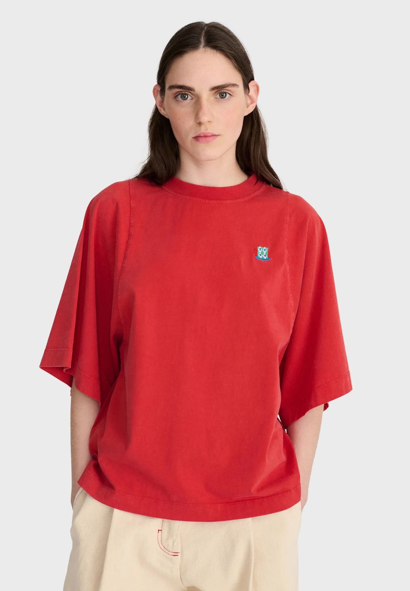 Personne aux longs cheveux foncés portant une chemise rouge ample avec un petit logo coloré sur la poitrine et un pantalon beige clair, debout sur fond blanc.