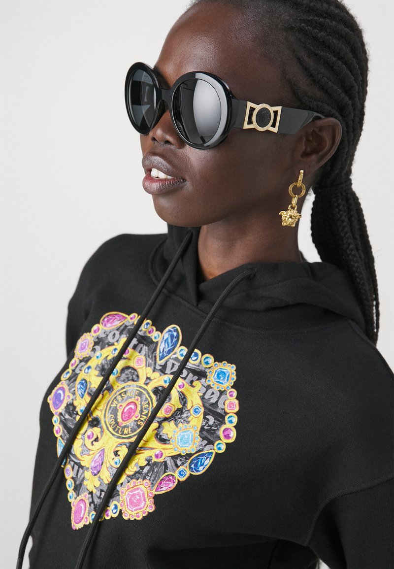 Un hoodie noir avec un design de cœur coloré présentant des accents bleus, roses et jaunes. De grandes lunettes de soleil noires et des boucles d'oreilles en or complètent le look.