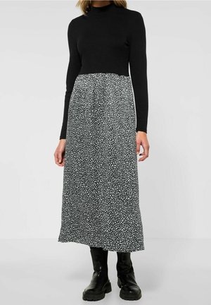 Frau trägt ein schwarzes Langarmshirt und einen schwarz-weiß gemusterten Midirock mit schwarzen Stiefeletten, steht vor einem schlichten Hintergrund.