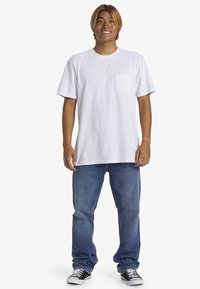 Camiseta de algodón blanca con mangas cortas y un bolsillo en el pecho, combinada con jeans de mezclilla azules y zapatillas negras y blancas.