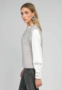 Débardeur gris tricoté sans manches avec un motif texturé, associé à une chemise blanche à manches longues avec des poignets en dentelle.