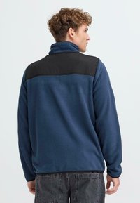 Sudadera de forro polar en azul marino con un panel superior negro, cuenta con un cuello alto y mangas largas, textura suave y corte casual.