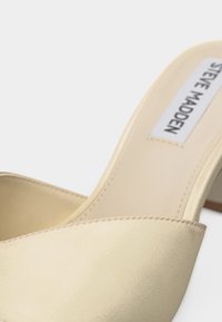 Mule beige à bout ouvert avec un petit talon et l'étiquette "Steve Madden" sur la semelle intérieure, sur un fond neutre.