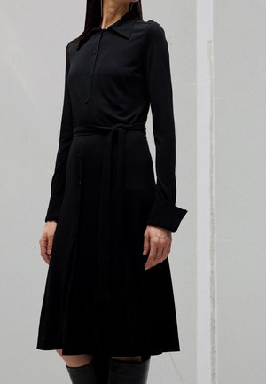 Femme portant une robe chemise noire à manches longues avec des boutons, une ceinture nouée à la taille, et des bottes noires montantes, sur un fond uni.