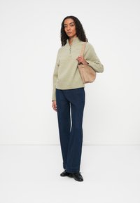 Suéter verde claro con cuello de cremallera, combinado con jeans de pernera ancha en azul marino. El modelo sostiene un bolso de hombro beige. Calzado negro completa el look.