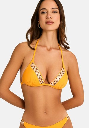 Femme portant un bikini triangle jaune avec une bordure à motifs, debout, les bras dans le dos, sur un fond clair uni.
