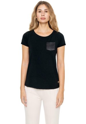 Une femme porte un tee-shirt noir à manches courtes avec une poche grise sur la poitrine et un pantalon blanc, sur un fond blanc uni.