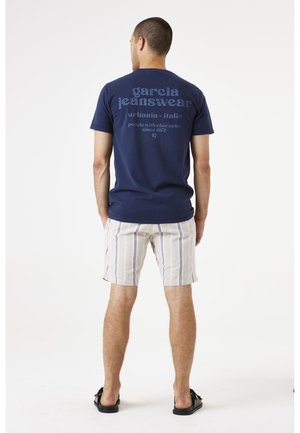 Navy blue cotton t-shirt with text on the back; paired with light-colored striped shorts; black flip-flops. Neutral background.