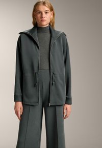 Tmavo šedá zipsová bunda s vysokým golierom, sťahovacím pásom a prednými vreckami, doplnená o zodpovedajúce široké nohavice a rebrovaný rolák.