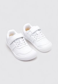 Zapatillas deportivas blancas con una textura suave, que cuentan con una correa de velcro y detalles de cordones. Punta redondeada y perforaciones en la parte superior.