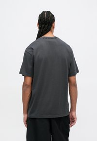 Hombre con el cabello trenzado largo, vistiendo una camiseta gris oscura y pantalones negros, de pie de espaldas contra un fondo blanco liso.