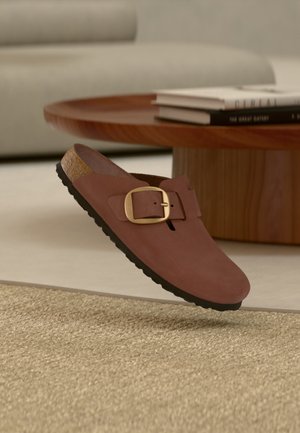 Καφέ παπούτσι slip-on με μεγάλο χρυσό αγκράφα, φελλό σόλα και μαύρη σόλα να αιωρείται πάνω από μπεζ χαλί κοντά σε ξύλινο τραπέζι με στοιβαγμένα βιβλία.