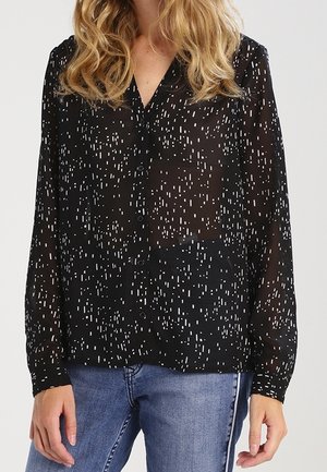 Femme portant un chemisier noir transparent à boutons avec un motif à pois blancs et un jean bleu, debout avec les mains détendues le long du corps.
