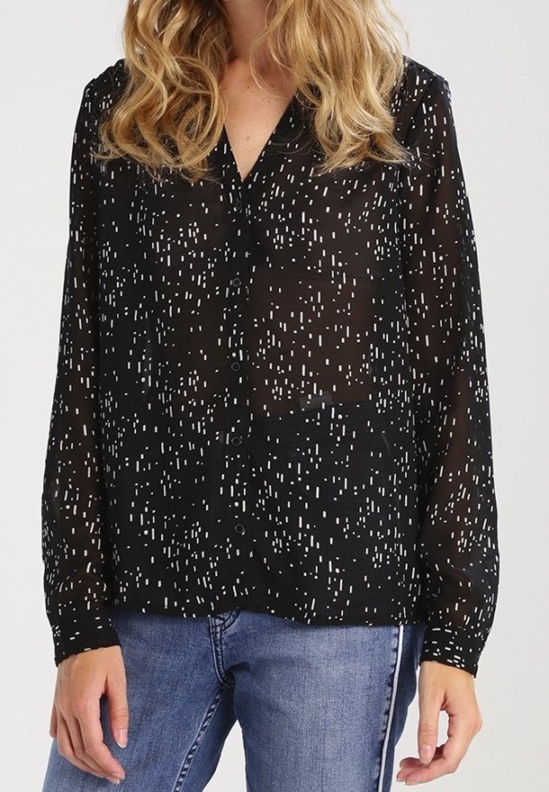 Femme portant un chemisier noir transparent à boutons avec un motif à pois blancs et un jean bleu, debout avec les mains détendues le long du corps.