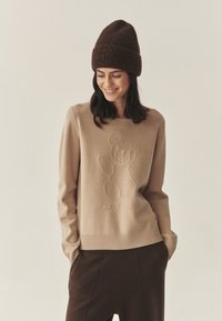 Maglione beige con design in rilievo di un orsetto, colletto e polsini a costine. Abbinato a pantaloni marroni scuri e un berretto di lana marrone.