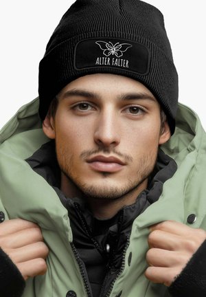 Black ribbed beanie with "ALTER FALTER" label; model in green puffer jacket. Black shirt underneath, showcasing layered textures and colors.