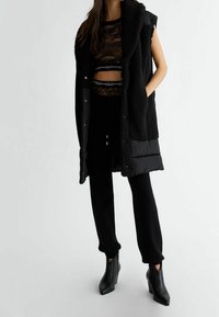 Czarna bezrękawnik o pluszowej fakturze, z kołnierzem szalowym i zapięciem na zatrzask; nałożony na przezroczysty czarny koronkowy top oraz dopasowane czarne spodnie.