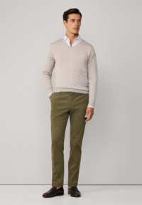 Mężczyzna w beżowym swetrze w serek na białej koszuli, w oliwkowych spodniach i ciemno-brązowych mokasynach, stojący na tle jasnego, jednolitego tła.