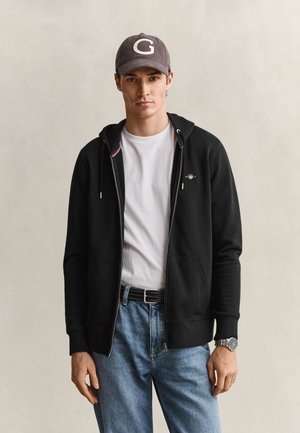 Jeune homme portant un sweat à capuche zippé noir, un t-shirt blanc, un jean bleu, une casquette grise avec la lettre G et une montre-bracelet argentée sur un fond uni.