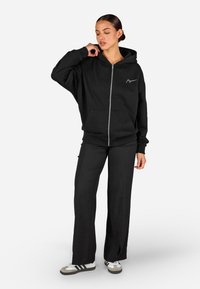 Fekete, cipzáras kapucnis pulóver kenguruzsebbel, oversize fazonban és bordázott mandzsettával, fekete nadrággal, oldalsó hasítékokkal párosítva. Fehér logó részlet.