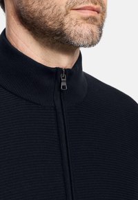 Nahaufnahme eines unteren Gesichts und Halses eines Mannes, der einen schwarzen texturierten Reißverschluss-Pullover trägt, dessen Reißverschluss teilweise geschlossen ist.