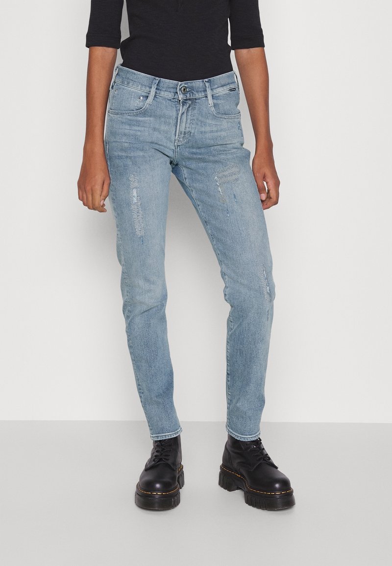 Jeans in denim blu chiaro con vestibilità slim, caratterizzati da toppe strappate e un classico design a cinque tasche. Abbinati a stivali neri con lacci.