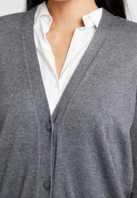 Cárdigan gris con escote en V, textura acanalada y tres botones. Lleva una camisa blanca de cuello debajo.