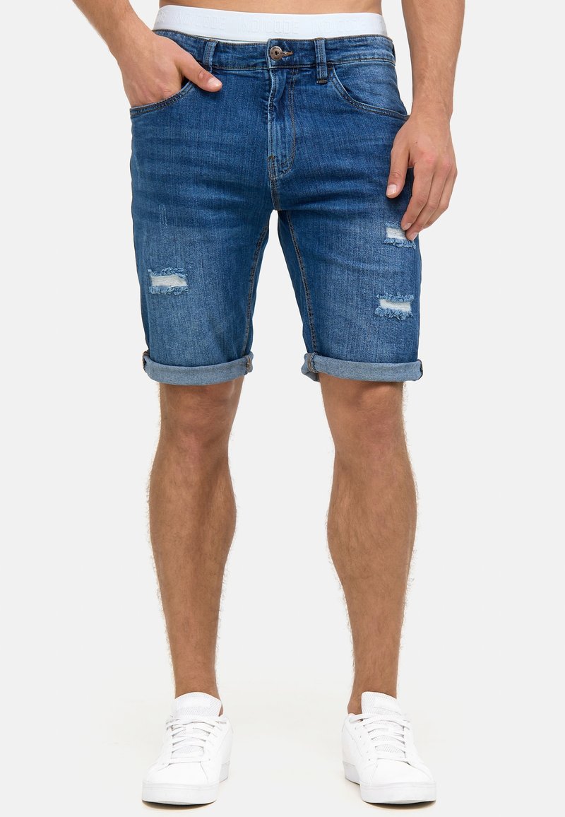 Shorts en denim bleu moyen avec des détails déchirés et des revers, portés avec des baskets blanches, sur un fond neutre.