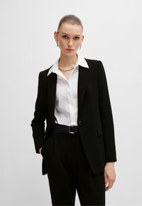 Czarna marynarka o dopasowanym kroju z jednym guzikiem, zestawiona z białą koszulą z kołnierzykiem oraz eleganckimi czarnymi spodniami. Uzupełniona łańcuszkiem i paskiem.