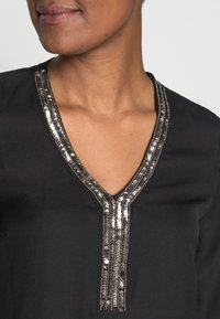 Schwarze Bluse mit V-Ausschnitt und dekorativem silbernem Paillettenbesatz in der Mitte, glatter Textur und tailliertem Schnitt.