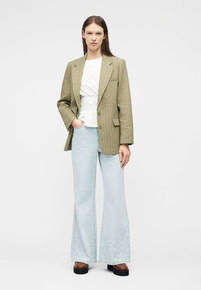 Femme debout portant un blazer rayé vert olive, un chemisier blanc, un jean large bleu clair avec un détail œillet, et des chaussures compensées marron.