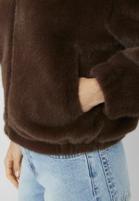 Veste en fausse fourrure marron à coupe décontractée, avec une texture douce, des poches latérales et des poignets côtelés. Associée à un jean denim bleu clair.