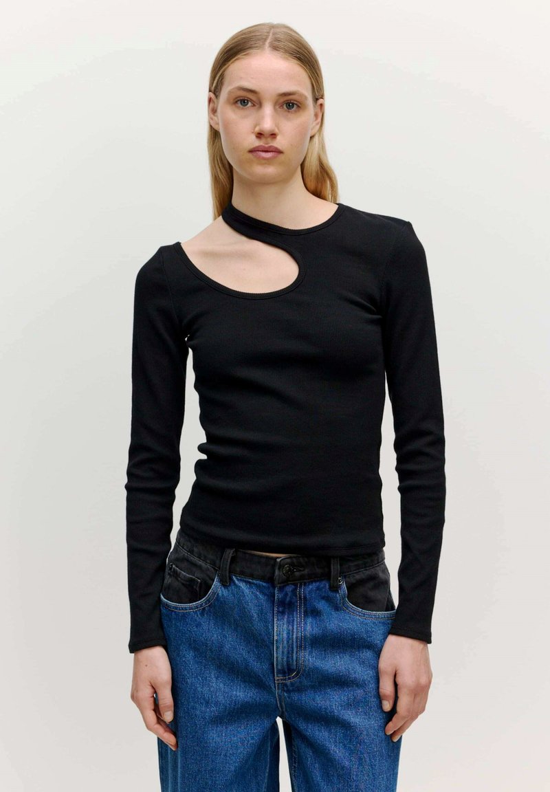 Schwarzes Langarmoberteil mit einem einzigartigen asymmetrischen Ausschnitt, aus geripptem Stoff. Getragen mit blauen hochtaillierten Jeans.