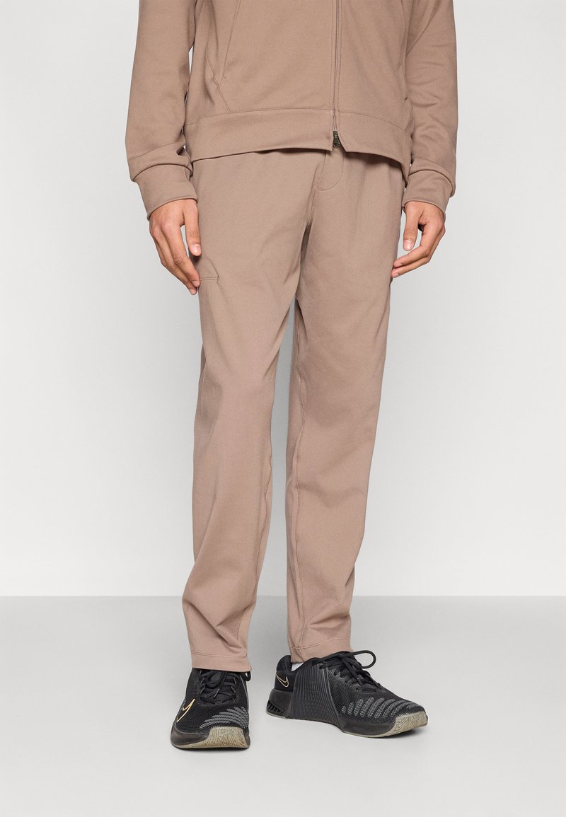 Καφέ αθλητικό παντελόνι με ομαλή υφή, εφαρμοστή γραμμή και πλαϊνή τσέπη, σε συνδυασμό με μαύρα παπούτσια με επάνω μέρος από δίχτυ και χρυσή λεπτομέρεια.