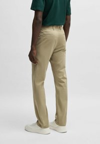 Chinos de algodón beige con corte recto, que presentan trabillas para cinturón, bolsillos traseros y una textura suave, combinados con zapatillas blancas.