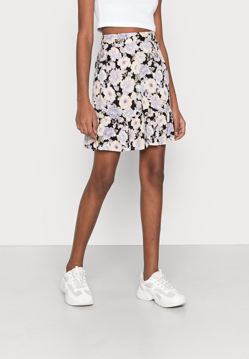 Femme portant une mini-jupe noire à fleurs pastel, un haut court blanc et des baskets blanches, debout sur un sol gris.