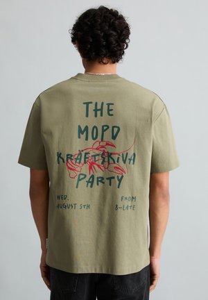 Persoon met krullend haar die een olijfgroen T-shirt draagt met een rode kreeft en tekst die reclame maakt voor "The Mopo Kräftskiva Party, woensdag 5 augustus, van 8 uur tot laat."