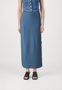 Jupe maxi en tricot côtelé bleu avec une taille ajust�ée. Portée avec des mules à bout ouvert noires, sur un fond blanc uni.