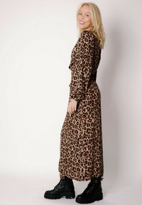 Beżowo-brązowa sukienka maxi w panterkę z miękkiego materiału, z długimi rękawami i talią podkreśloną paskiem, zestawiona z masywnymi czarnymi botkami.
