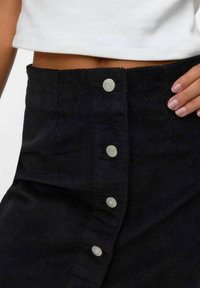 Jupe en velours côtelé noir avec une surface texturée. Présente de grands boutons blancs le long de la fermeture à l'avant et un design taille haute.