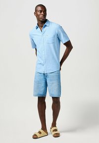 Jasnoniebieska koszula na krótki rękaw z guzikami, z jedną kieszenią na piersi, w połączeniu z jasnymi dżinsowymi szortami i beżowymi sandałami wsuwanymi.