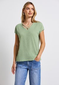 Top verde chiaro a V con maniche corte, caratterizzato da un tessuto testurizzato e spalline incrociate all'icollo. Abbinato a jeans in denim blu chiaro.
