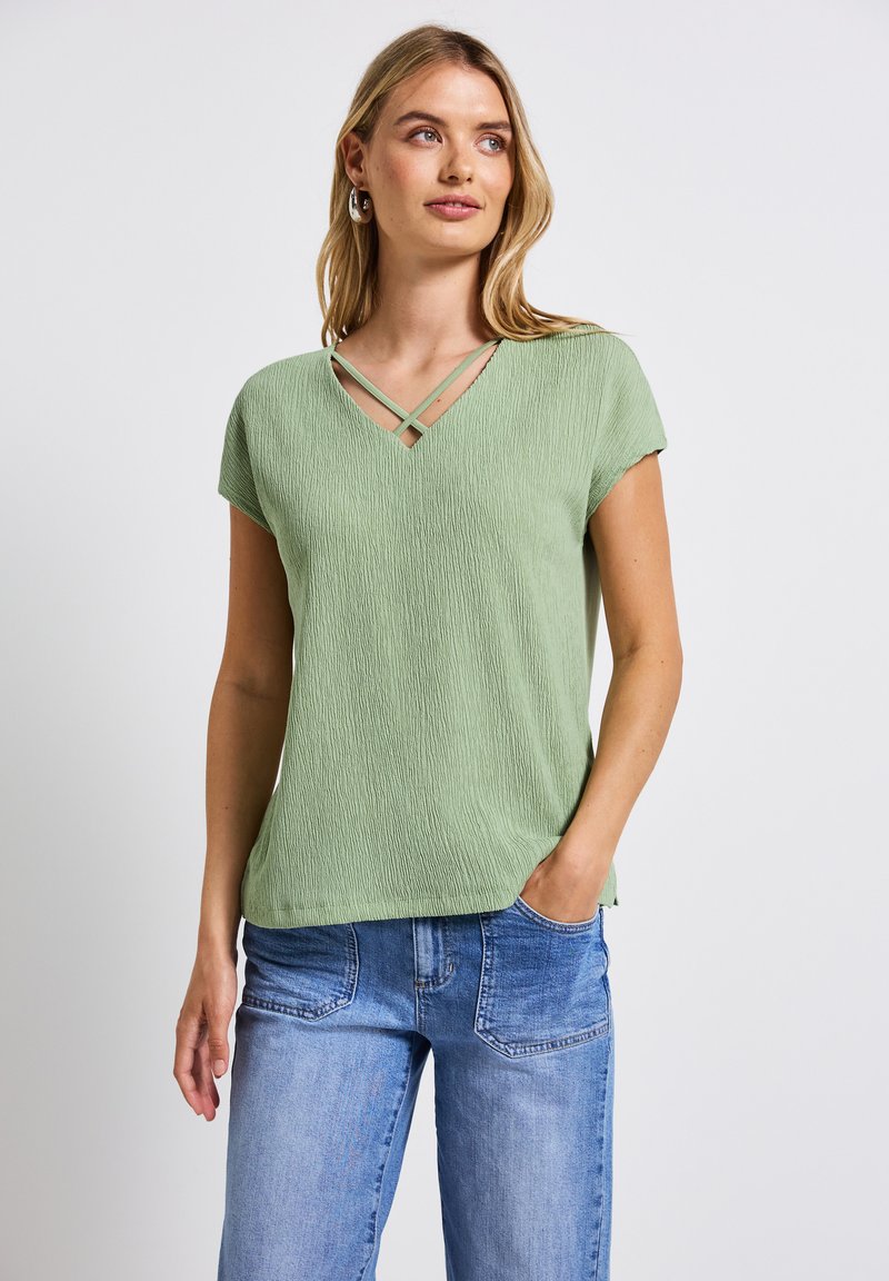 Top verde chiaro a V con maniche corte, caratterizzato da un tessuto testurizzato e spalline incrociate all'icollo. Abbinato a jeans in denim blu chiaro.