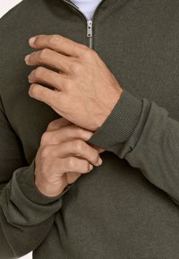 Donkergroen gebreide pullover met een ritssluiting aan de kraag, voorzien van geribbelde manchetten. Handen die de mouw aanpassen tonen fijne textuur en naaddetails.