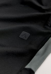 A fekete atlétikai anyag sima textúrájú, finom zöld szegéllyel és egy fekete háromszög alakú logóval ellátva.