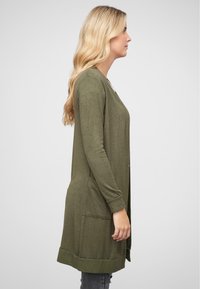 Vihreä oliivinvärinen pitkä neule, joka on valmistettu pehmeästä kankaasta. Siinä on suora leikkaus, avonainen etuosa ja kaksi sivutaskua. Ribbaus hihansuissa ja helmassa.