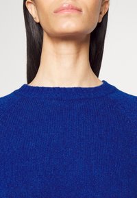 Modrý pletený svetr s kulatým výstřihem a ribovanou texturou, s raglánovými rukávy a jemným povrchem. Detailní pohled na oblast límce.