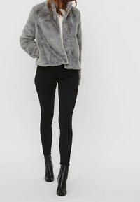 Šedá kožešinová bunda s volným střihem, předními kapsami a vysokým límcem, v kombinaci s černými skinny kalhotami a kotníkovými botami.