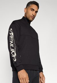 Black zip-up sweatshirt made of textured fabric, featuring a contrasting logo design in beige on the left sleeve and ribbed cuffs.