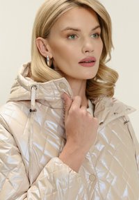 Donna con capelli biondi corti che indossa una giacca beige trapuntata e lucida con cappuccio, mentre tiene il colletto vicino al viso, guardando di lato.